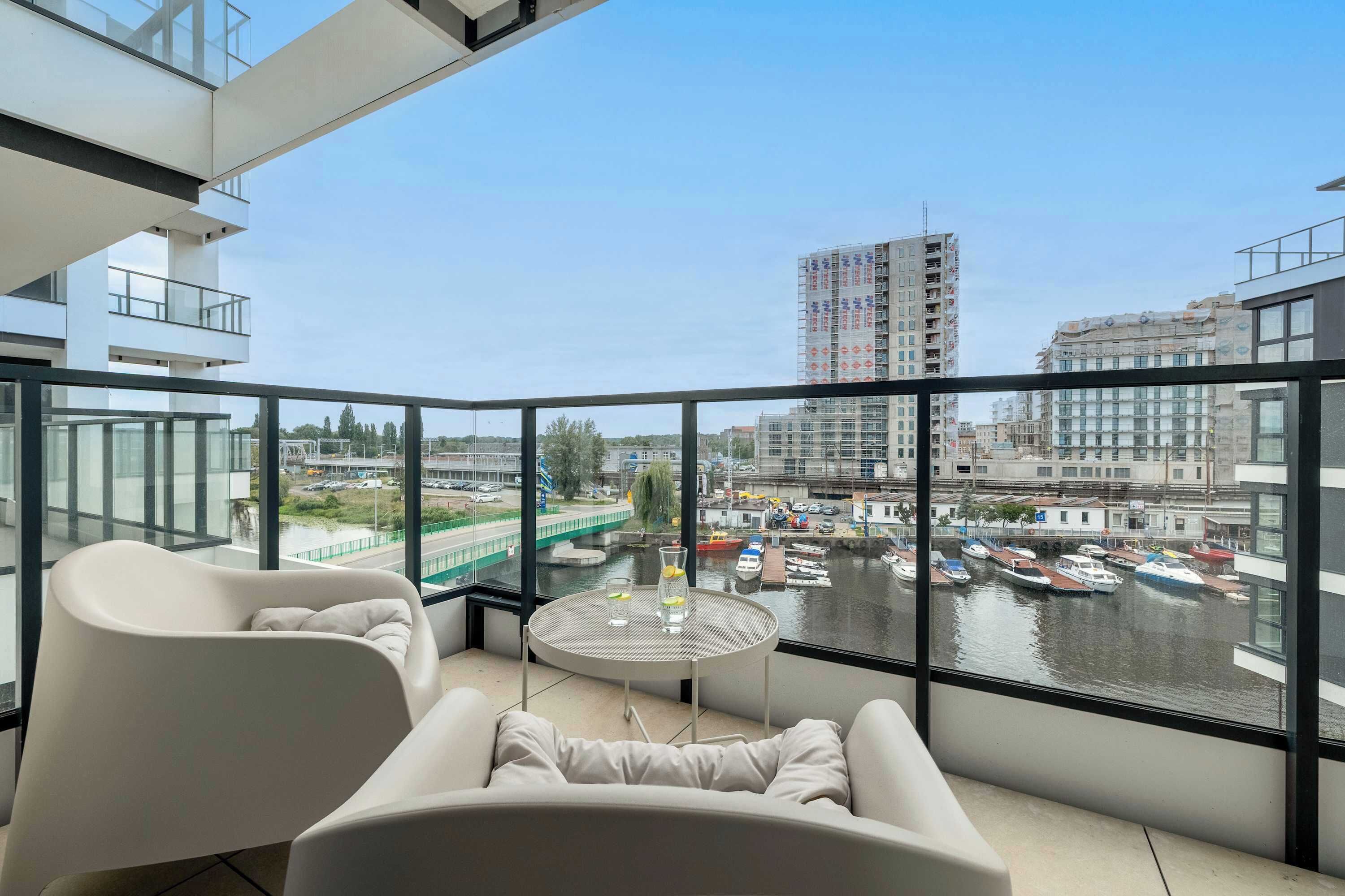 Un balcón moderno con vistas a un río y barcos, barandilla de vidrio y zona de estar con mesa.