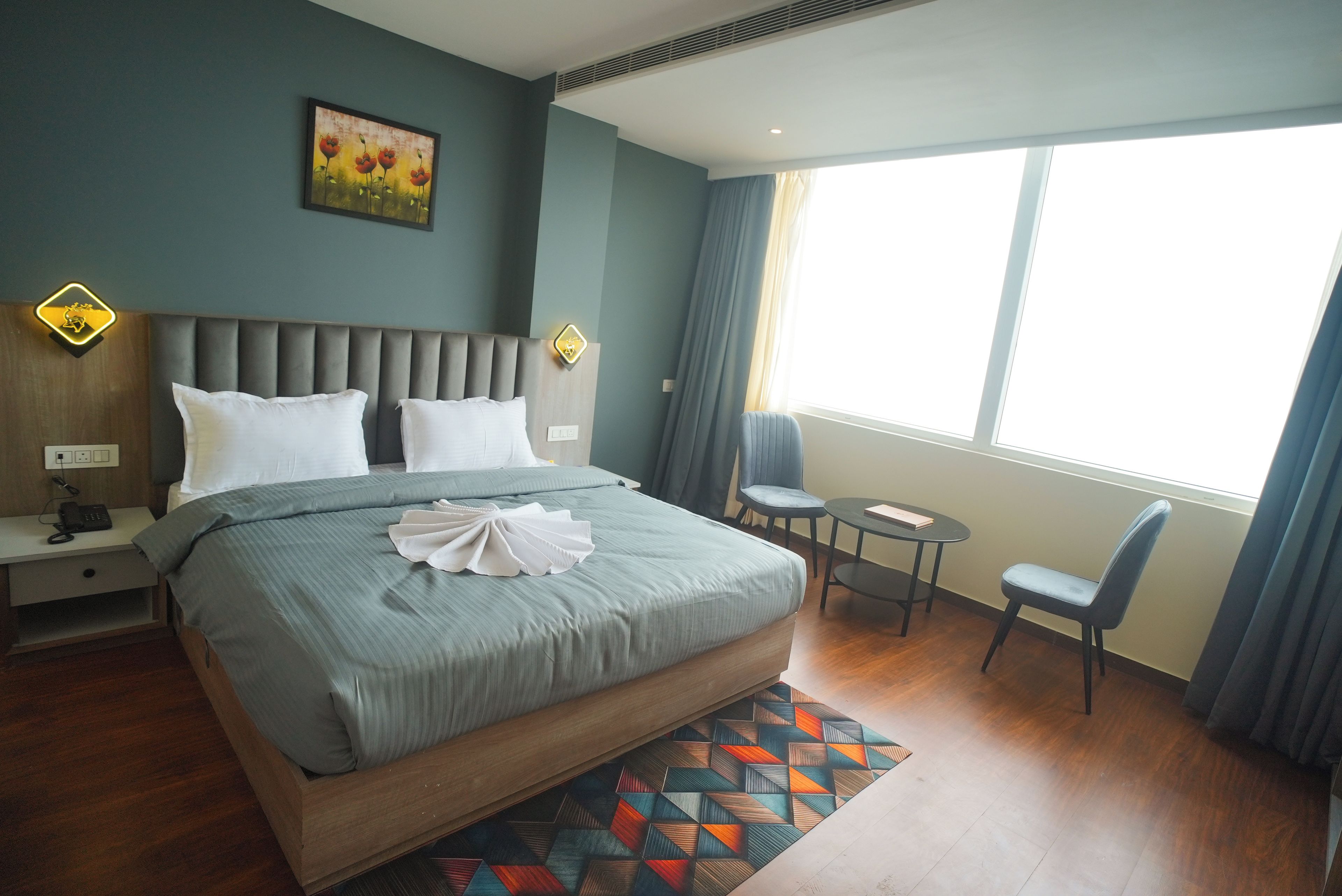 A hotel room with a large bed, a bedside table with a phone, a chair, a small table, and a window with curtains.