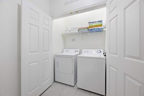 A laundry room with two white washers and dryers, a shelf with folded towels, and a white door. - The Happily Ever After Hideaway (Kissimmee)