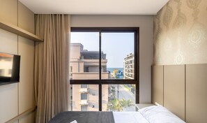 A hotel room with a bed, a TV, a large window with a view of buildings and a street, and a patterned wall. - Apt sea View, Pool Condo, 300m to Beach Csd0302 (Bombinhas)