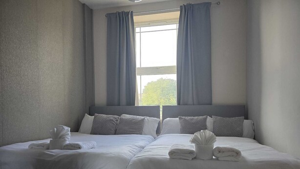 A double bed with grey bedding and pillows, a bedside table with a cup, and a window with curtains. - Cosy Flat near Canary Wharf | Sleeps 6 (London)