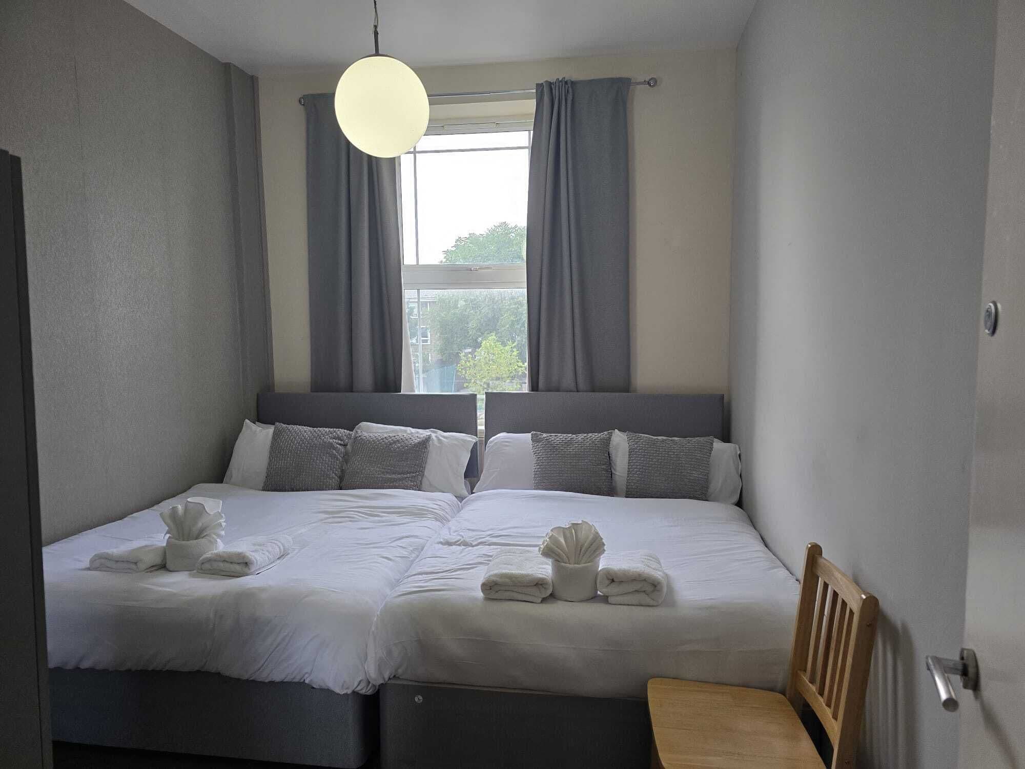 A double bed with grey bedding and pillows, a bedside table, a wooden chair, a hanging light fixture, and a window with curtains.