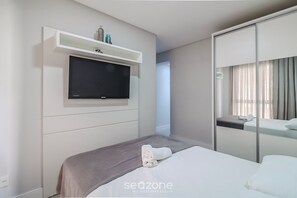 Ein modernes Hotelzimmer mit einem Bett, einem an der Wand befestigten Fernseher, einem Nachttisch mit Handtuch und einem Spiegel mit Blick aus dem Fenster.