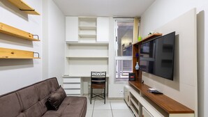 A compact living space with a sofa, a TV, a desk with a chair, and built-in shelving. - 1BR Apart With Self Check-in, Asa Sul Gdp0010 (Brasília)
