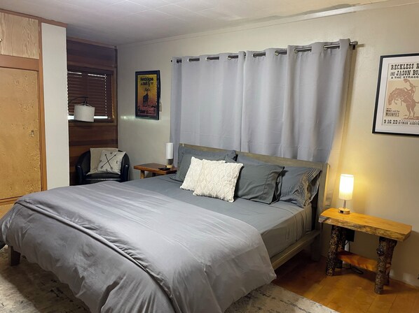 Une chambre avec un grand lit, une table de chevet avec une lampe, une commode en bois, une chaise et une affiche accrochée au mur.