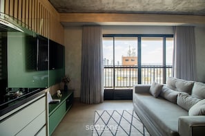 A modern living room with a sofa, a television, and a view of the cityscape through large windows. - Studio in a Condominium With a Pool Idvb2103 (Goiânia)