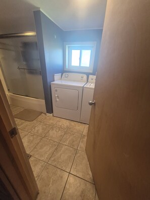 A laundry room with a washing machine and dryer, a window above the dryer, and a door leading to another room.