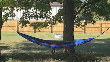 A hammock hanging between two trees in a grassy yard with a wooden fence in the background.