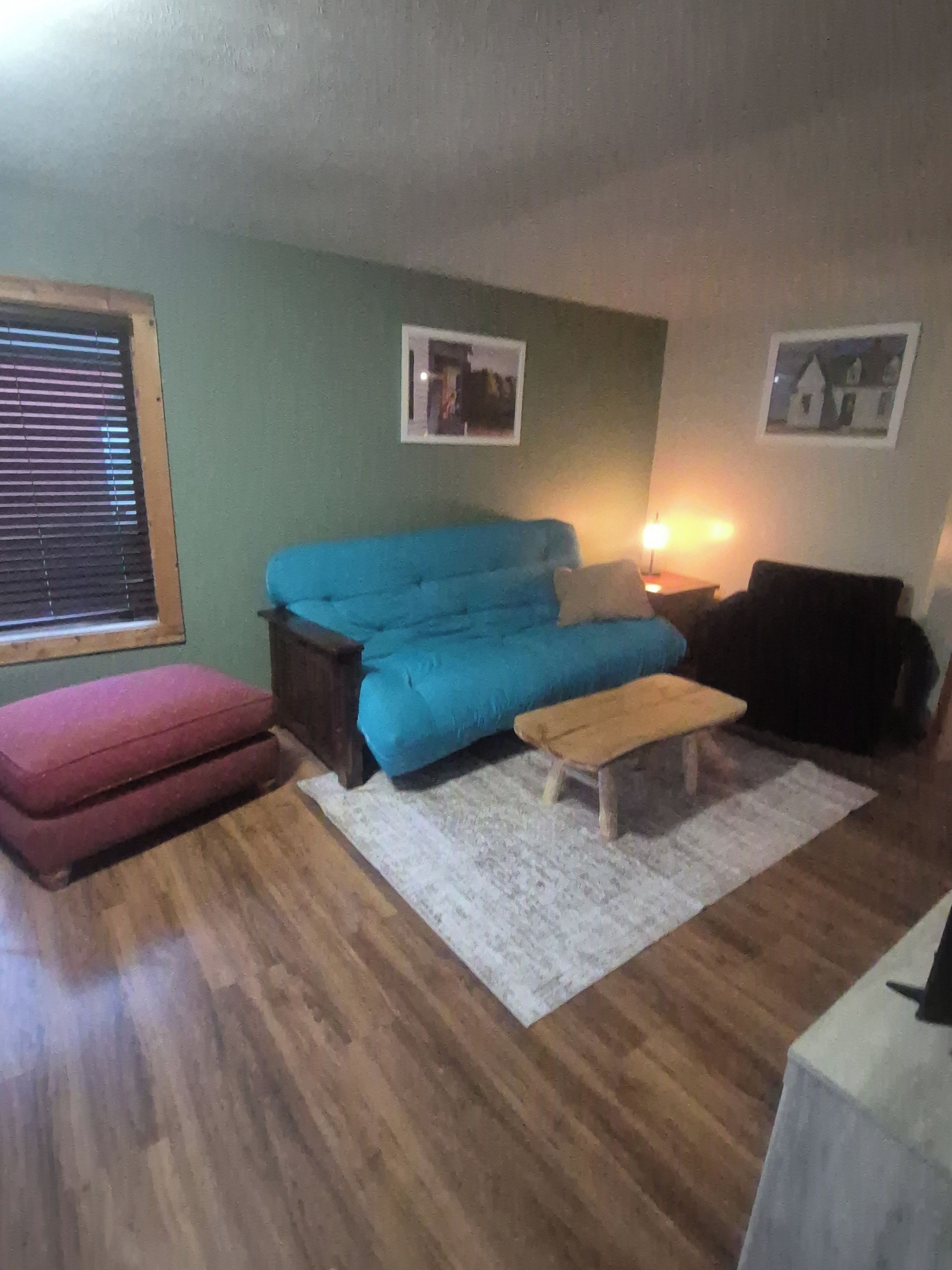 Een woonkamer met een blauwe bank, een rode poef, een houten bankje en een wit vloerkleed. Twee ingelijste foto's hangen aan de muur.