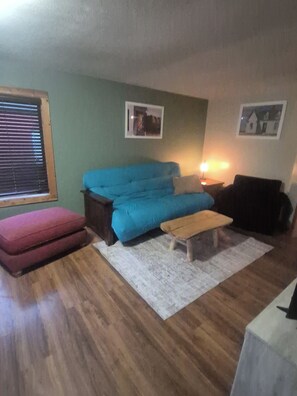A living room with a blue sofa, a red ottoman, a wooden bench, and a white rug. There are two framed pictures on the wall. - Peaceful 2-bedroom Private Cabin on 1 Acre with Hot Tub. (Greenville)