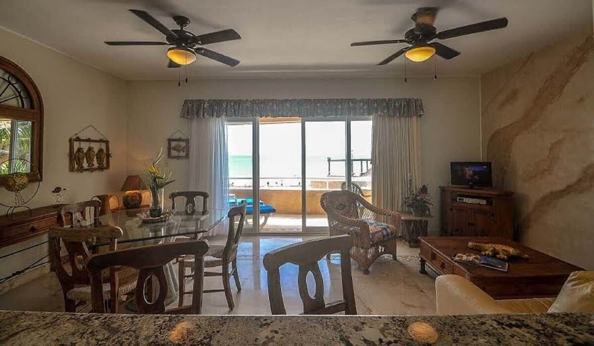 A living room with a glass dining table, two ceiling fans, a television, and a view of the beach.