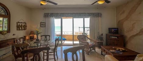 Un salon avec une table à manger en verre, deux ventilateurs de plafond, un téléviseur et une vue sur la plage.