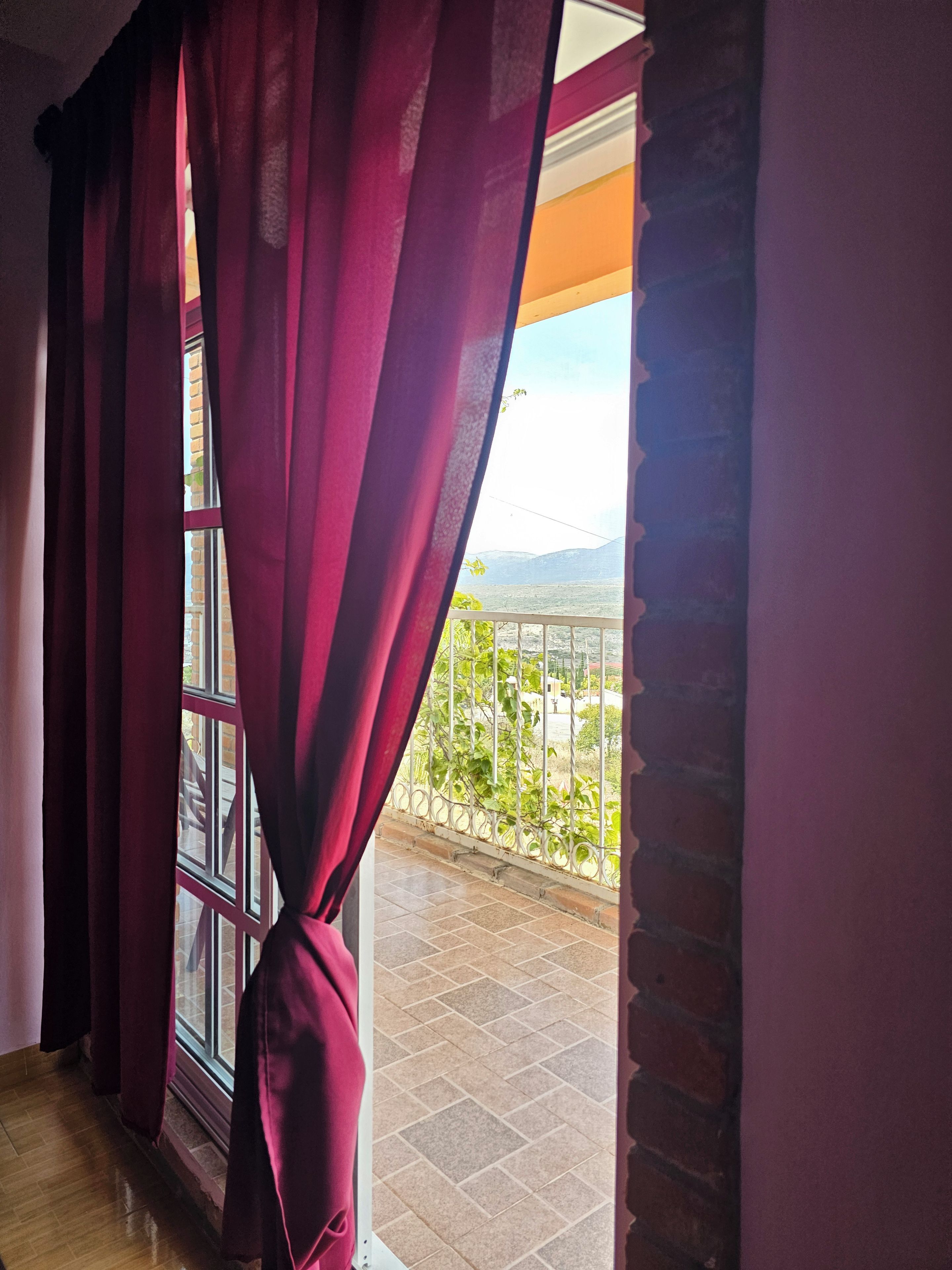 Una habitación con ventana cubierta por cortinas rojas, un balcón con barandilla de metal y vista a un paisaje lejano.