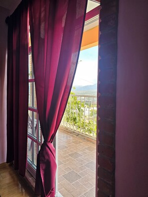 A room with a window covered by red curtains, a balcony with a metal railing, and a view of a distant landscape.