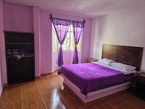 Un dormitorio con una cama, cabecera de madera, un armario y una ventana con cortinas moradas.