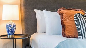 Een net opgemaakt bed met witte en gestreepte kussens, een grijze hoofdbord en een blauw-witte lamp op een klein tafeltje ernaast.