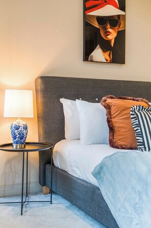 Ein ordentlich bezogenes Bett mit weißen und gestreiften Kissen, einem grauen Kopfteil und einer blau-weißen Lampe mit Muster auf einem kleinen Nachttisch daneben.