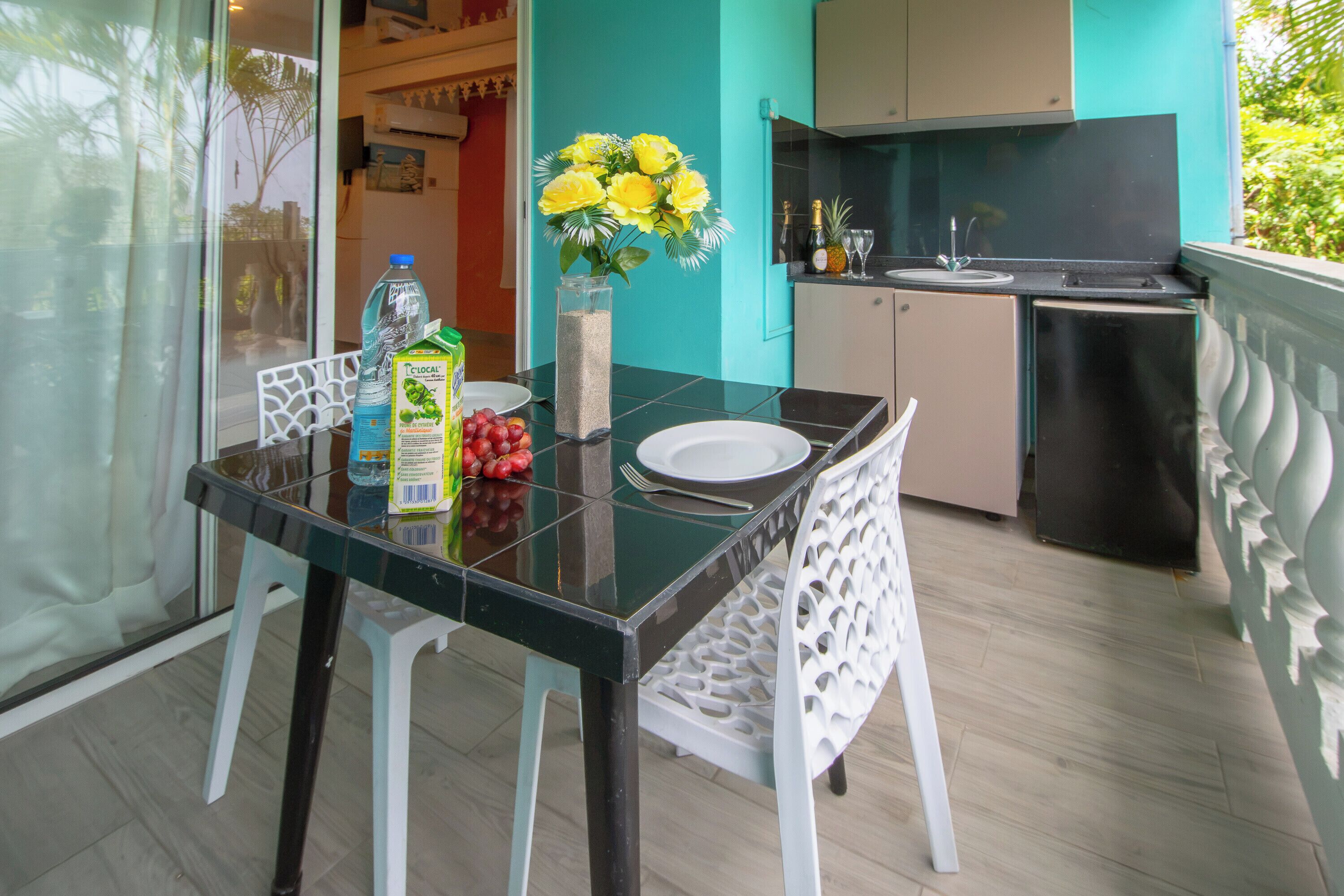 A dining area with a glass table, white chairs, a vase with yellow flowers, a bottle of water, and a carton of juice.