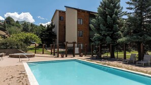 A swimming pool with a wooden deck, lounge chairs, and a building with a brick facade. - Lofted Mountain Lookout (Steamboat Springs)