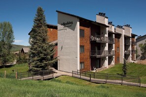 Um edifício de vários andares com varandas, uma placa com a inscrição 'The Rockies Condominiums' e uma área gramada com um deck de madeira.