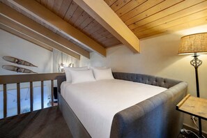 Ein Schlafzimmer mit einem großen Bett, Holzbalkendecke, einem Balkon mit Geländer und einer Lampe auf einem Nachttisch.