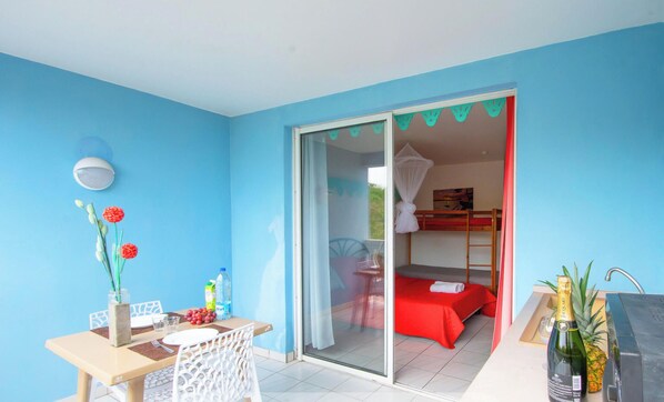 A room with a sliding glass door leading to a bedroom with a bed and a red blanket. There is a small table with a vase of flowers and a bottle of water. - Rêve Bleu 14 - Clim - Proche plage (LE DIAMANT)