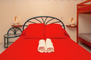 A single bed with red bedding and two pillows, a glass side table, a wooden nightstand with a shell decoration, and a bunk bed to the right. - Rêve Bleu 14 - Clim - Proche plage (LE DIAMANT)