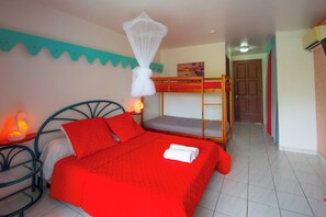 Ein Zimmer mit Etagenbett, einem Einzelbett mit roter Bettwäsche, einem Moskitonetz, einem Bild an der Wand und einer Tür im Hintergrund.