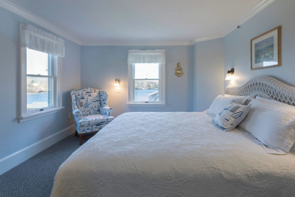 A bedroom with a large bed, two windows with curtains, a chair, and wall-mounted lights.
