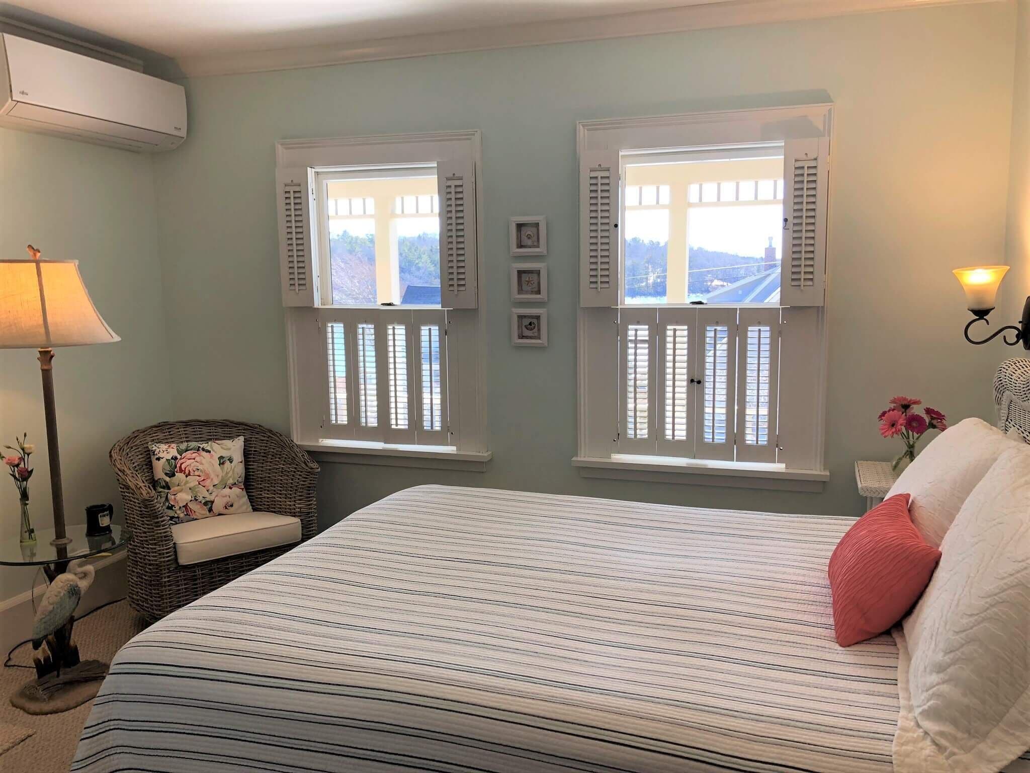 A bedroom with a bed, two windows with shutters, a lamp, a chair, and a wall-mounted light fixture.