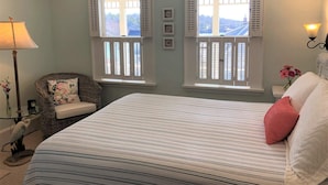 A bedroom with a bed, two windows with shutters, a lamp, a chair, and a wall-mounted light fixture. - Five Gables Inn (East Boothbay)