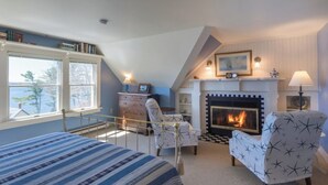 A bedroom with a bed, two chairs, a fireplace, and a window overlooking a body of water. - Five Gables Inn (East Boothbay)