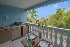 A balcony with a dining table set for two, a kitchen area, and a view of the surrounding greenery. - Rêve Bleu 5 - Clim - Proche plage (LE DIAMANT)