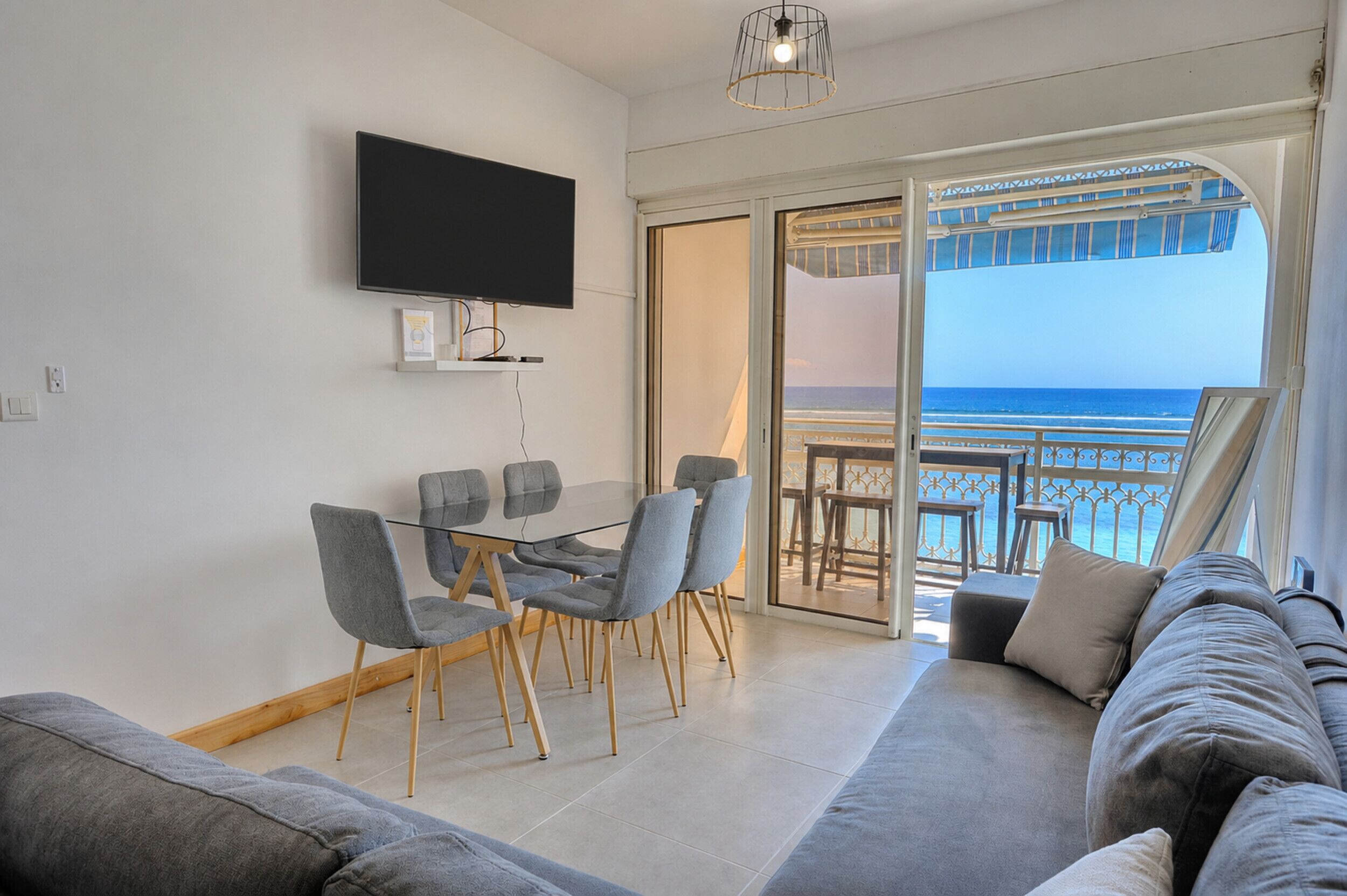 Un moderno salón con televisor de pantalla plana, una mesa de vidrio con sillas, un sofá y un balcón con vistas al mar.