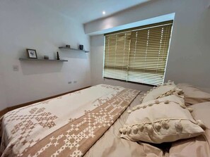 Un dormitorio con cama, ventana con persianas, estante con objetos y una colcha estampada.