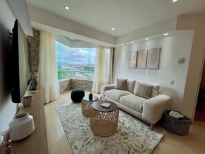 A modern living room with a sofa, two round coffee tables, a flat-screen TV, and a large window offering a city view. - Vistas Increibles y Confort en El Rosal (Caracas)