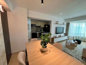 Ein modernes Wohnzimmer mit Esstisch, Stühlen, einer Couch, einem Fernseher und einer Küche im Hintergrund.
