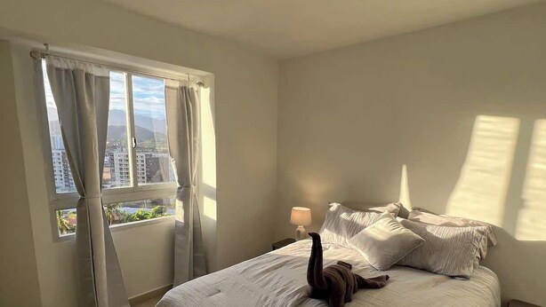 Un dormitorio con una cama grande, una mesita de noche, una lámpara y vistas a edificios por la ventana.