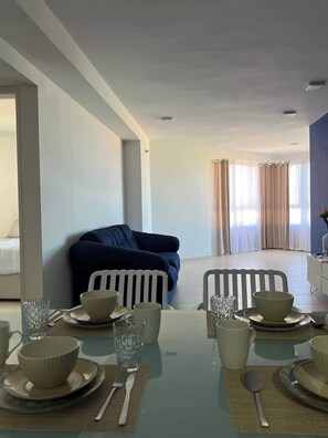 Een eethoek met een tafel voor twee personen, een blauwe bank op de achtergrond en uitzicht op een kamer met gordijnen.