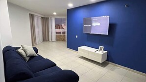 Una sala moderna con un sofá azul oscuro, un mueble bajo para televisor blanco y un ventanal con cortinas.