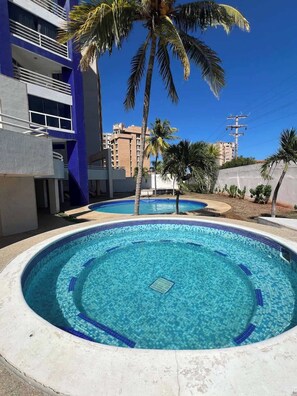 Ein Swimmingpool mit Fliesenrand, umgeben von Palmen und einem Gebäude mit Balkonen.