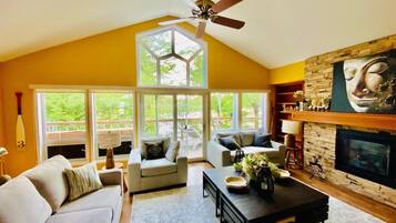 A spacious living room with a high ceiling, a fireplace, and a large window overlooking a deck.