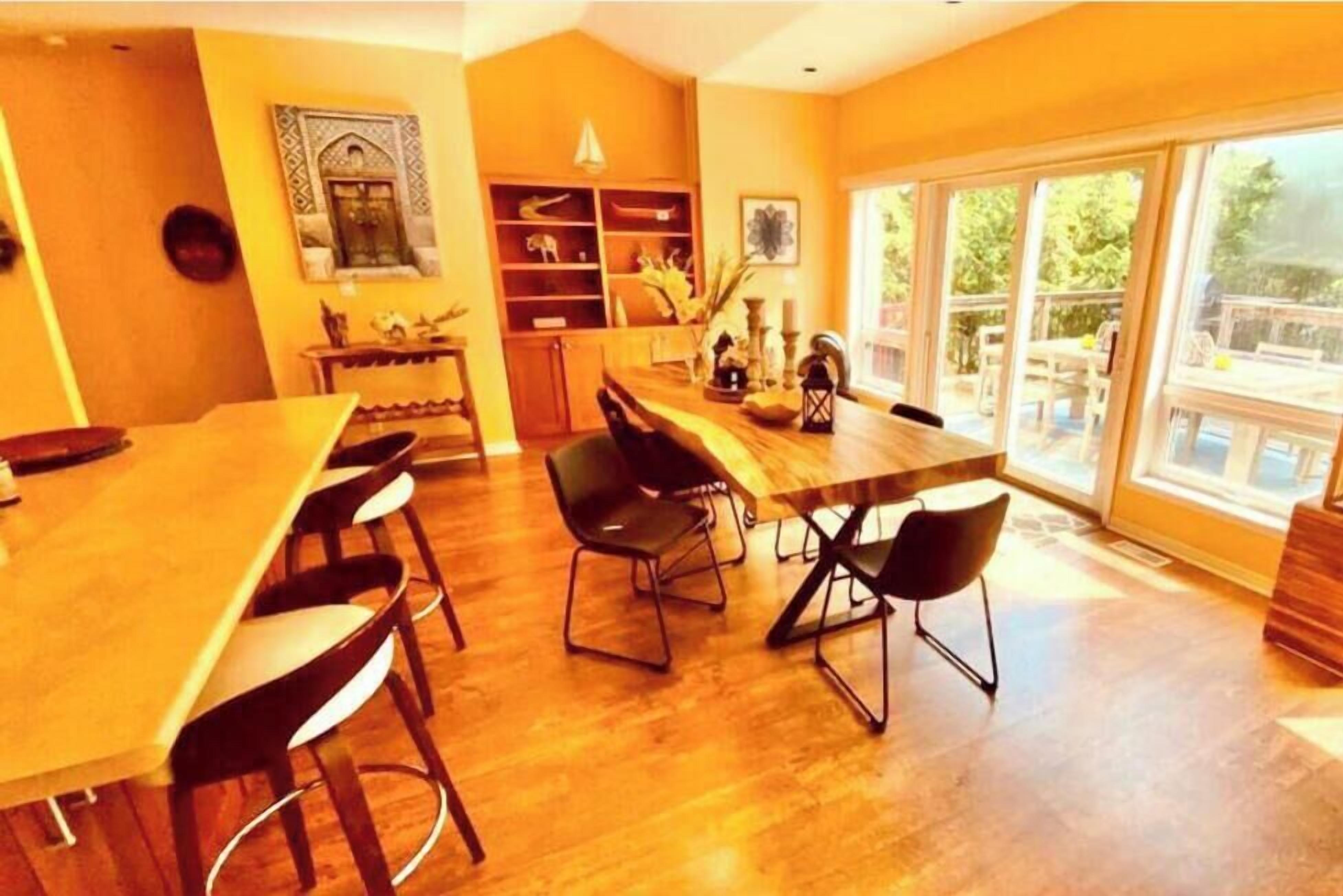 Un espace repas avec une longue table et des chaises en bois, une bibliothèque et une vue sur l’extérieur grâce à de grandes fenêtres.