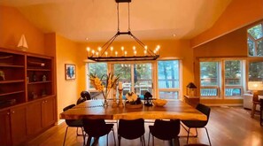 Un espace repas avec une grande table en bois, des chaises noires, un lustre et de grandes fenêtres.