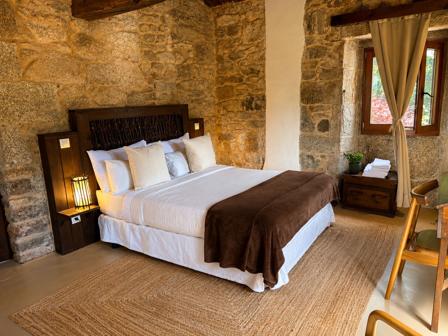 Un dormitorio con pared de piedra, cabecero de madera, cama con ropa de cama blanca, manta marrón, mesita de noche con lámpara, una silla de madera y una ventana con cortinas.