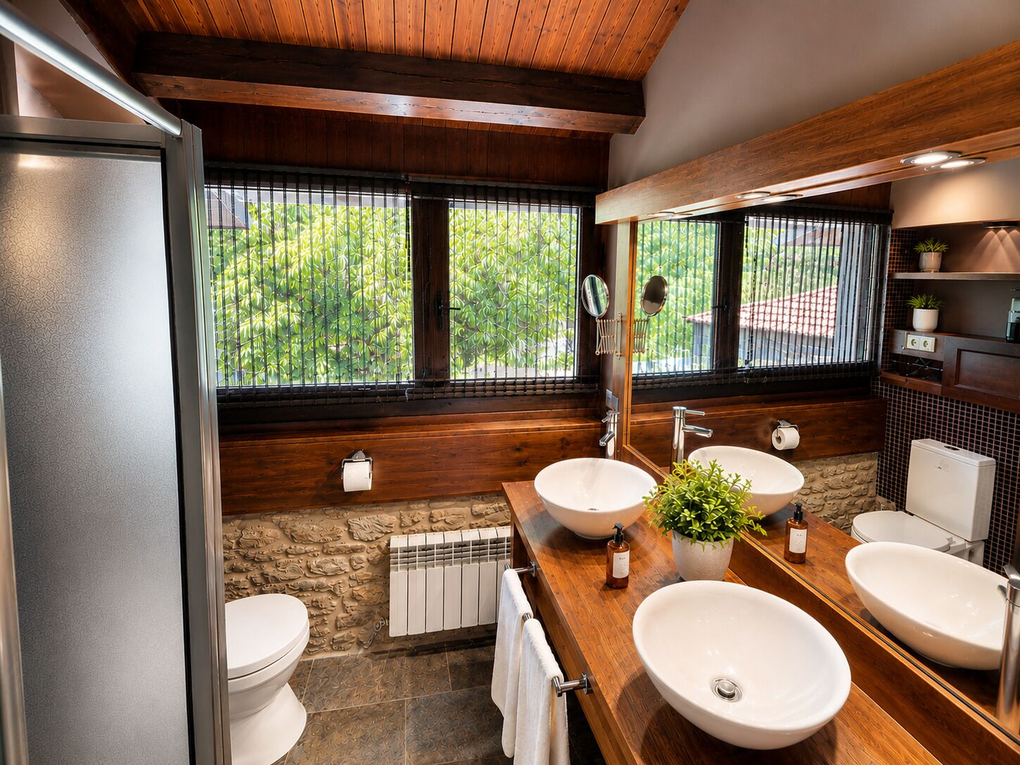 Et moderne badeværelse med træloft, to håndvaske, et toilet og et stort vindue med udsigt til grønne områder.