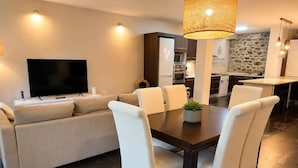 A modern living room with a sofa, dining table, and chairs, a flat-screen TV, and a kitchen area in the background.