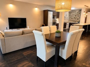 A modern living room with a sofa, dining table, and chairs, a flat-screen TV, and a kitchen area in the background.