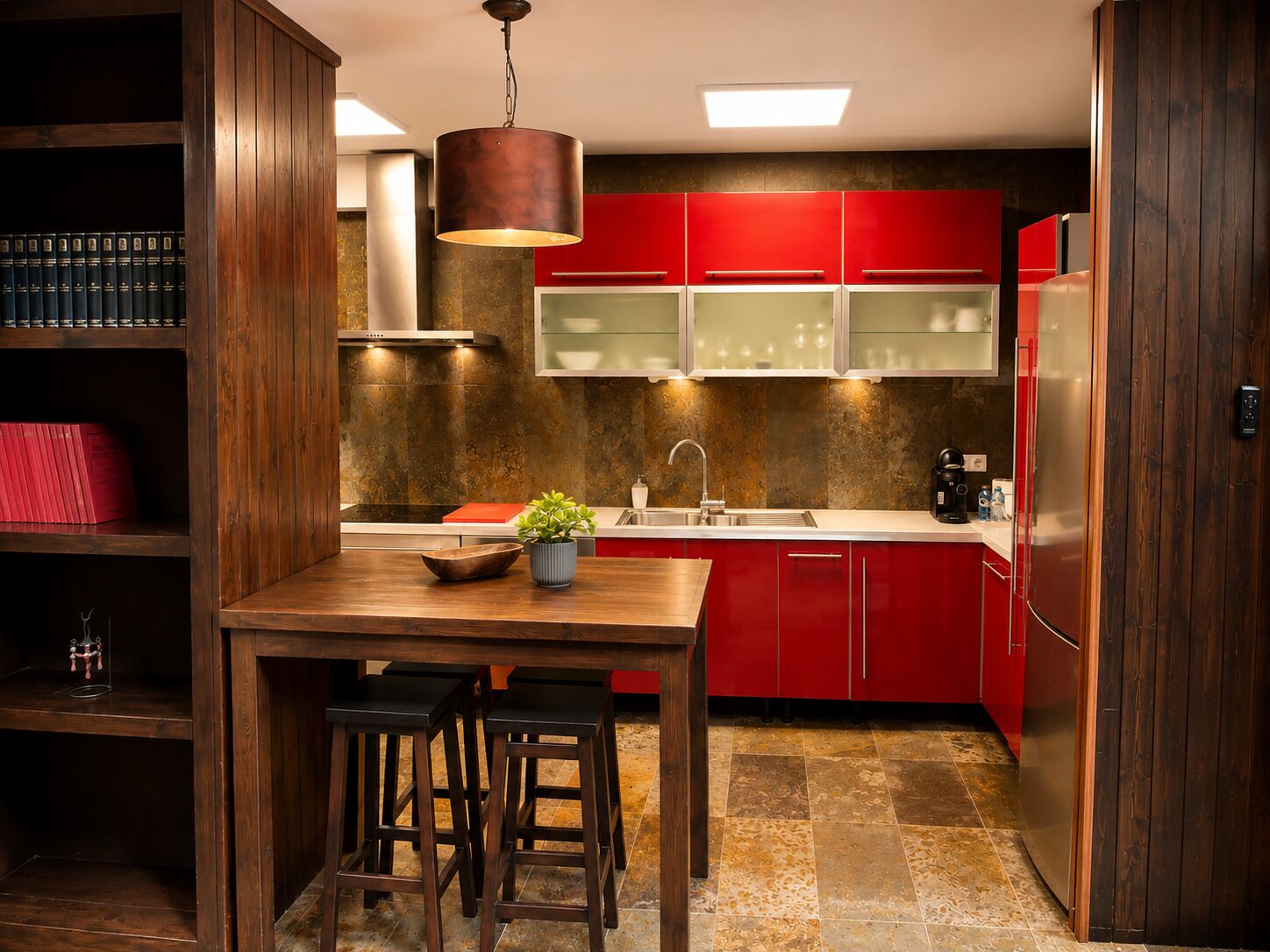 Una cucina con mobili rossi, un tavolo di legno con sgabelli, una lampada a sospensione e una libreria a parete.