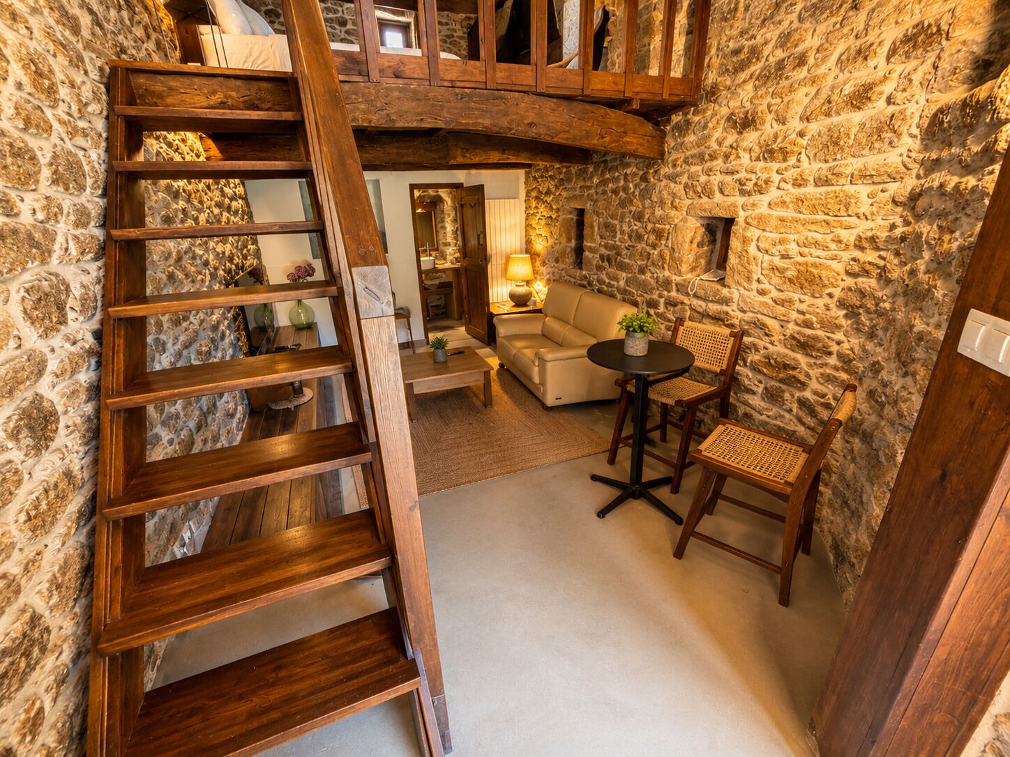 Un interior rústico con una escalera de madera, paredes de piedra, un sofá y una mesa con sillas.
